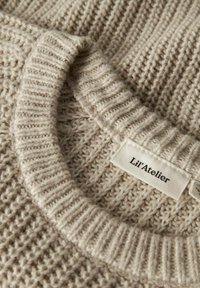 Šviesiai smėlinis megztinis su tekstūruotu raštu ir kraštu pagal apykaklę. Etiketei užrašyta "Lil'Atelier." Minkšta, jauki medžiaga.