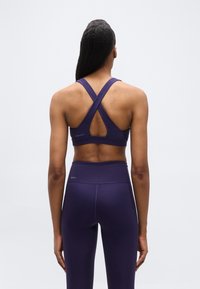 Γυναίκα με μακριά μαλλιά, φορώντας σκούρο μωβ αθλητικό μπουστάκι με σταυρωτή πλάτη και κολάν με ψηλή μέση, στέκεται με την πλάτη στραμμένη προς τα εμπρός σε απλό φόντο.