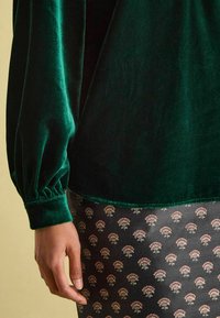 Dunkelgrünes Samtoberteil mit langen Ärmeln und gerafften Manschetten; kombiniert mit einem gemusterten Rock, der kleine florale Motive auf schwarzem Hintergrund zeigt.