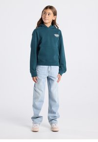 Teal hoodie with a front pocket and a logo, paired with light blue jeans and beige sneakers. Hoodie features a drawstring and ribbed cuffs.