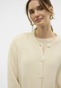 Beige Strickjacke mit rundem Halsausschnitt, ausgestattet mit zwei Knöpfen und einem strukturierten Stoff. Leicht und locker sitzendes Design.