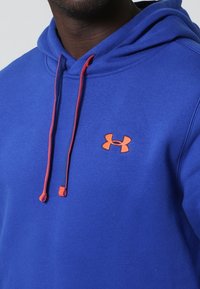 Niebieska bluz sweatshirtowa z kapturem, z pomarańczowymi sznurkami i logo Under Armour na piersi. Materiał wydaje się być miękki o gładkiej fakturze.