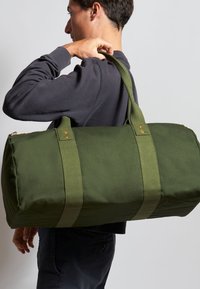 Grøn kanvas duffeltaske med robust stof og dobbelt skulderremme, har gyldent hardware og en rund form.
