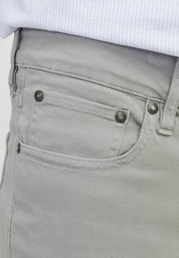 Šedé džíny z denimu s boční kapsou a kovovými nýty, hladkou texturou a dvojitě šitými švy, částečně zakryté světle modrou pruhovanou košilí.