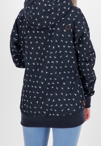 Marinblå hoodie med vitt fågelmönster, bärs av person i ljusblå jeans, sedd bakifrån mot en enfärgad bakgrund.