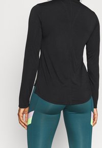 Schwarzes langärmliges Sportoberteil mit abgerundetem Saum und Nahtdetails, kombiniert mit türkisfarbenen Leggings mit mehrfarbigen geometrischen Mustern.