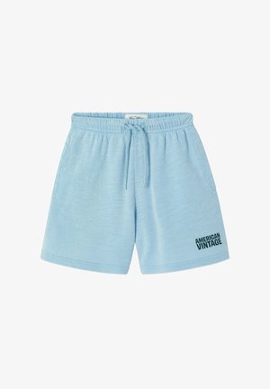 Blond dziecko stojące, ubrane w granatowy t-shirt, jasnoniebieskie szorty i brązowe klapki na jednolitym jasnym tle.