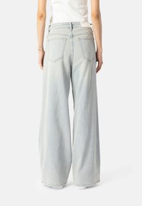 Osoba u svijetloplavim širokim trapericama i bijeloj majici, stoji s opuštenim rukama, prikazana sa leđa na jednobojnoj pozadini.