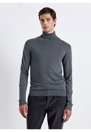 Giovane uomo con maglione grigio aderente a collo alto e pantaloni scuri, in piedi davanti a uno sfondo chiaro e uniforme.