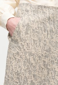 Main dans la poche d'une jupe taille haute avec un motif tissé texturé beige et gris, associée à un chemisier à manches longues de couleur crème.
