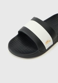 Svart og hvit skyvesandal med en teksturert svart såle, en bred øvre stropp med en hvit stripe og en liten gull logo aksent.
