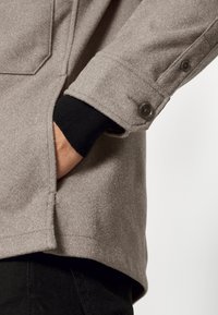 Persoon in een beige wollen jas met zwarte knopen, rechterhand in de zijzak gestoken, zwarte mouwen zichtbaar eronder.