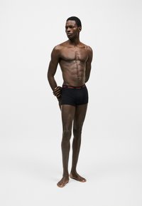 Homme athlétique debout pieds nus sur fond uni, portant un boxer noir avec une ceinture rouge "HUGO", les bras dans le dos, regardant sur le côté.