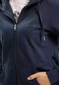 Granatowa bluza z kapturem na zamek, wykonana z miękkiego materiału, z kieszenią kangurzą i małym logo "Coco Jeans" na piersi.