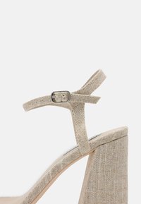 Beige hoge hak sandaal met een bovenkant van textuurstof, enkelband met een zilveren gesp en een chunky hak.
