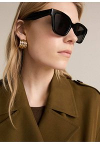 Gafas de sol negras de estilo angular, pendientes de espiral dorados y una blusa marrón que lleva una capa bajo un abrigo trench verde oliva con cuello y hombreras.