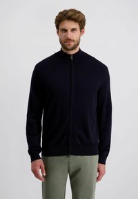 Marineblauer Reißverschluss-Pullover aus weichem Stoff, mit einem hohen Kragen, gerippten Bündchen und schmalem Schnitt. Kombiniert mit hellgrünen Hosen.