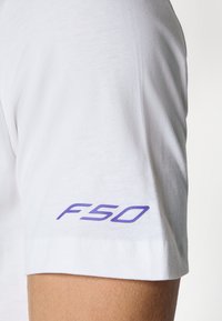 T-shirt blanc à manches courtes avec une texture lisse, présentant un "F50" violet imprimé sur la manche gauche. Design simple et épuré.