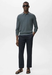 Gray knit polo sweater with a collar, navy trousers, and black sandals. Model wears sunglasses, arms relaxed at sides, standing on a plain background.