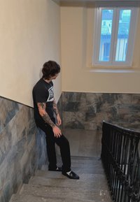 Schodisko so šedými kamennými schodmi, čiernym kovovým zábradlím a stenami z krémových a vzorovaných dlaždíc. Osoba stojí, oblečená v čiernom tričku a nohaviciach.