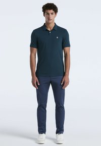 Ein junger Mann steht aufrecht und trägt ein dunkelblaues Poloshirt, eine marineblaue Hose und weiße Turnschuhe vor einem schlichten hellen Hintergrund.