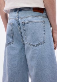 Άτομο που φοράει ανοιχτό μπλε τζιν με φαρδιά μπατζάκια και λευκό τοπ, εμφανιζόμενο από τη μέση πίσω μέχρι το μέσο του μηρού.