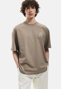 Brun bomulds t-shirt med en afslappet pasform, der har en rund halsudskæring og korte ærmer, trykt med "VI ER FREMTIDEN" i hvid.
