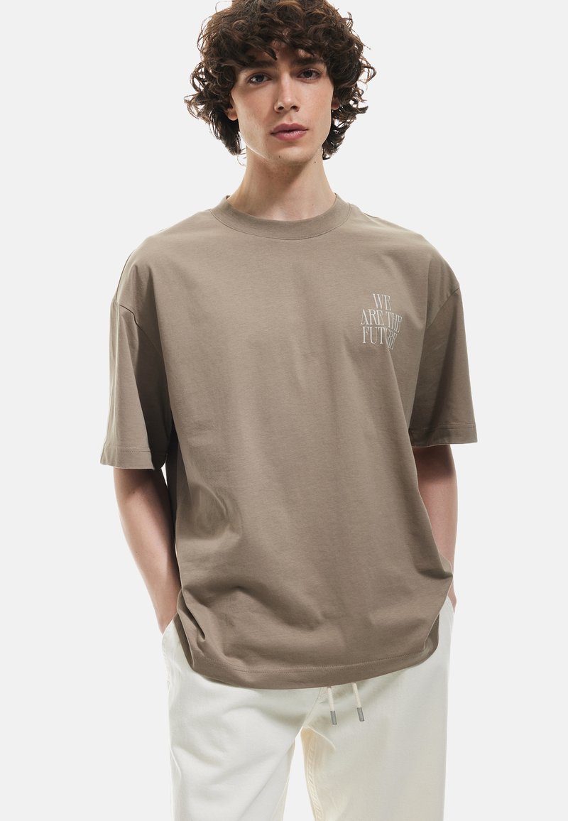 Brun bomulds t-shirt med en afslappet pasform, der har en rund halsudskæring og korte ærmer, trykt med "VI ER FREMTIDEN" i hvid.