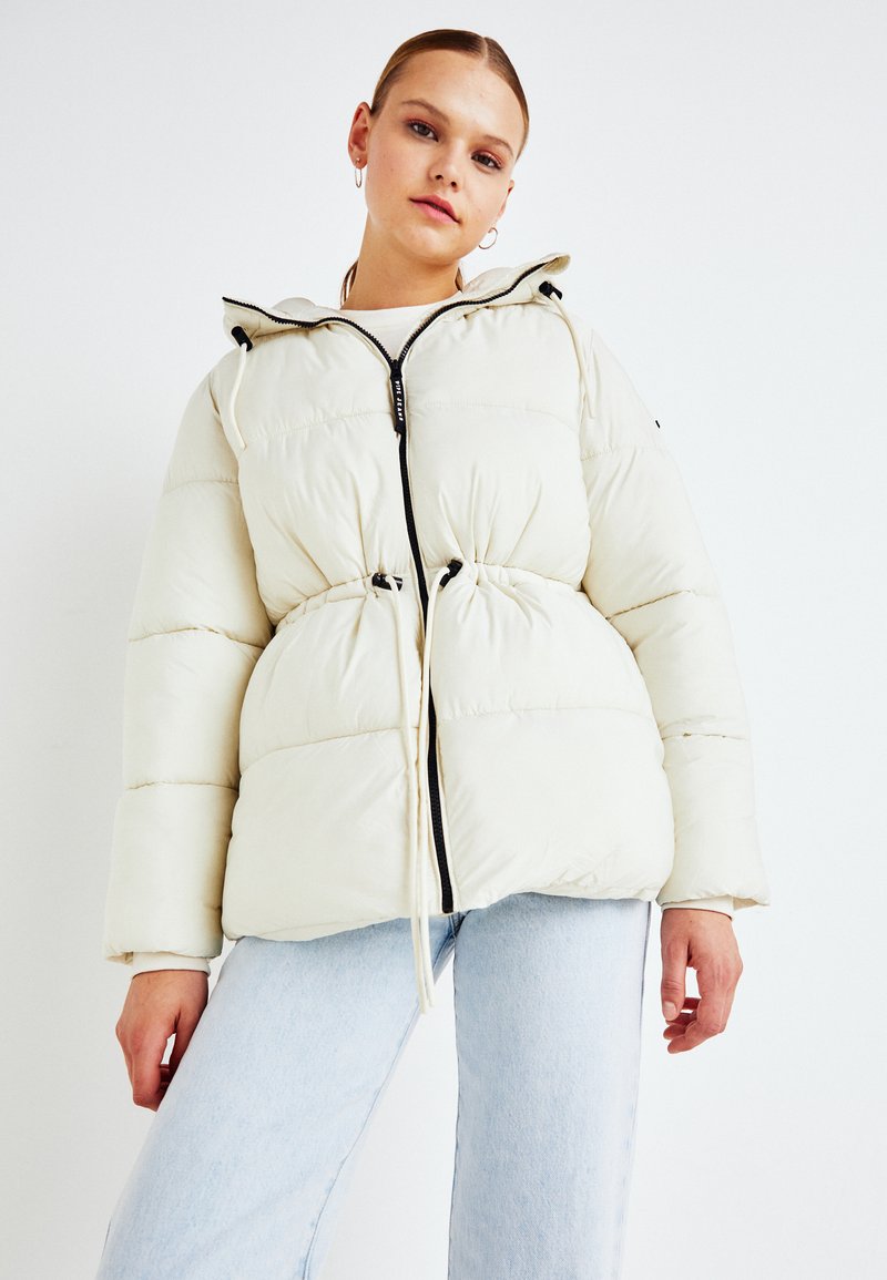 Chaqueta acolchada color crema con cintura ajustada, cremallera negra y capucha; presenta una textura suave y cordones elásticos. Combinada con jeans claros.