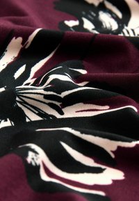 Kankaan kukkakuosi, jossa on viininpunainen tausta, mustia ja kermaisia kukkia. Tekstuuri näyttää sileältä ja pehmeältä.