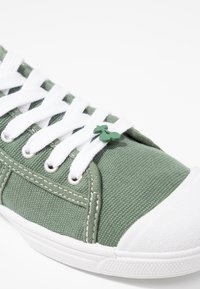 Sneaker in tela verde con punta in gomma bianca e lacci. Presenta un piccolo accento verde vicino ai lacci. Tessuto testurizzato visibile.