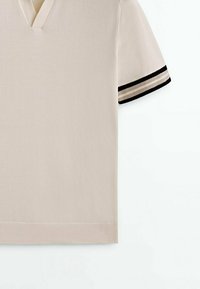 Polo en maille beige à manches courtes avec col en V et rayures noires et beiges aux poignets, présenté sur fond blanc.