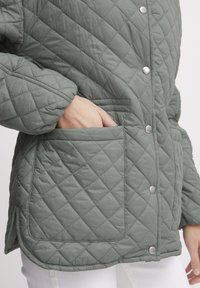 Padded žiemos striukė švelniai žaliai, su spaustukais, deimantine raštu ir šonine kišenė. Minkšta, paminkštinta medžiaga su lygiu paviršiumi.