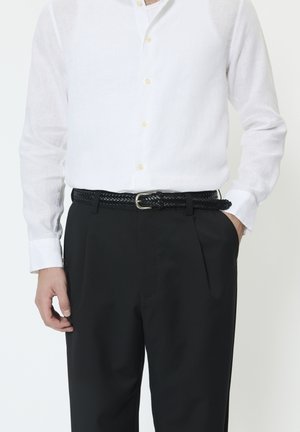 Schwarzer gewebter Gürtel mit silberner Schnalle, getragen über schwarzen Hosen, kombiniert mit einem weißen Leinenhemd mit Knopfleiste und Kragen.