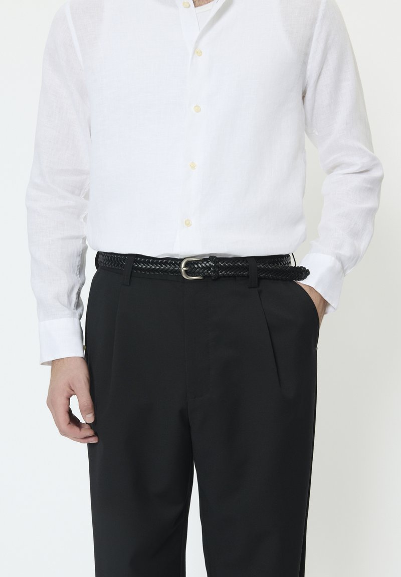 Curea neagră împletită cu o cataramă argintie purtată peste pantaloni negri, asortată cu o cămașă albă din in, cu nasturi și guler.