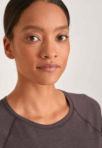 Graues Rundhalsoberteil mit Raglanärmeln, glatter Textur und dezenten Stichdetails. Das Model trägt ein natürliches Makeup und hat glattes Haar.