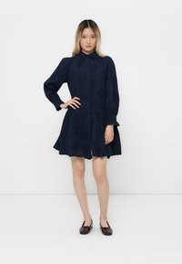 Woman with long ombre hair wearing a navy blue embroidered dress with long puffed sleeves and dark red ballet flats, standing against white wall.