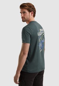 T-shirt med korte ærmer i teal med et grafisk design i crème og blå på ryggen, lavet af blød bomuldsblanding.
