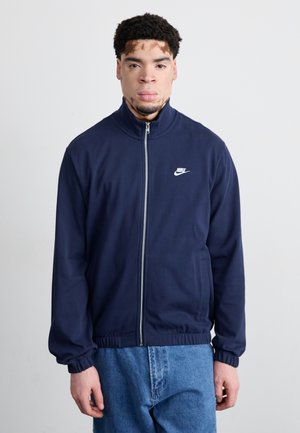 Marineblå zip-up jakke med høj krave, elastiske manchetter og elastisk talje. Har et lille hvidt Nike-logo på brystet. Bæres med blå jeans.