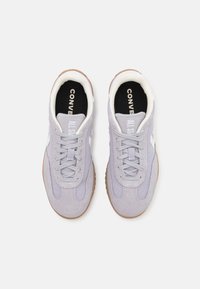 Pár světle šedých semišových a síťovaných tenisek s bílými tkaničkami, gumovými podrážkami a nápisem "Converse All Star" vytištěným uvnitř boty.