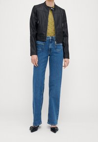 Chaqueta de moto de cuero negro con cuello de presión, blusa texturizada verde, jeans rectos azules y zapatos negros de punta afilada con detalles metálicos.