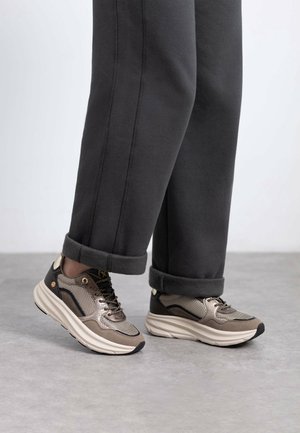 Personne portant un pantalon noir et des baskets beiges, noires et blanches, debout sur un sol gris.