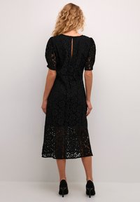 Black lace midi dress featuring short puff sleeves, floral pattern, keyhole back detail, and a slightly flared hem. Paired with black heels.