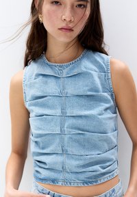 Ljusblå ärmlös denimtop med vikta texturer, rund halsringning och synliga sömmar. Bärs med matchande denimunderdelar.