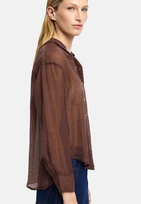 Brązowa, lekka, półprzezroczysta koszula zapinana na guziki z kołnierzykiem, długimi rękawami i lekko zaokrąglonym dołem, noszona na niebieskich dżinsach.