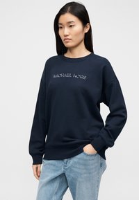 Σκούρο μπλε φόρμα με στρογγυλή λαιμόκοψη και μακριά μανίκια, με ραμμένο λογότυπο «MICHAEL KORS» σε λευκό στο στήθος, συνδυασμένο με ανοιχτό μπλε τζιν.