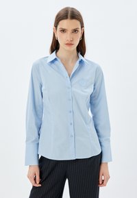 Camisa de botón azul claro hecha de una tela suave, con cuello puntiagudo, mangas largas y un logo bordado en el pecho.
