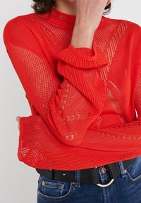 Red knitted sweater featuring a mesh pattern, long sleeves, and a ribbed neckline, layered over a black belt and denim jeans.