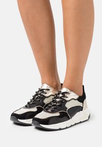 Černobílá teniska s kombinací materiálů, kovovými akcenty, texturovanými překryvy a masivní podrážkou s vlnitým vzorem.