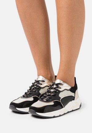 Schwarz-weißer Sneaker mit einer Mischung aus Materialien, metallischen Akzenten, strukturierten Überzügen und einer klobigen Sohle mit Wellendekor.